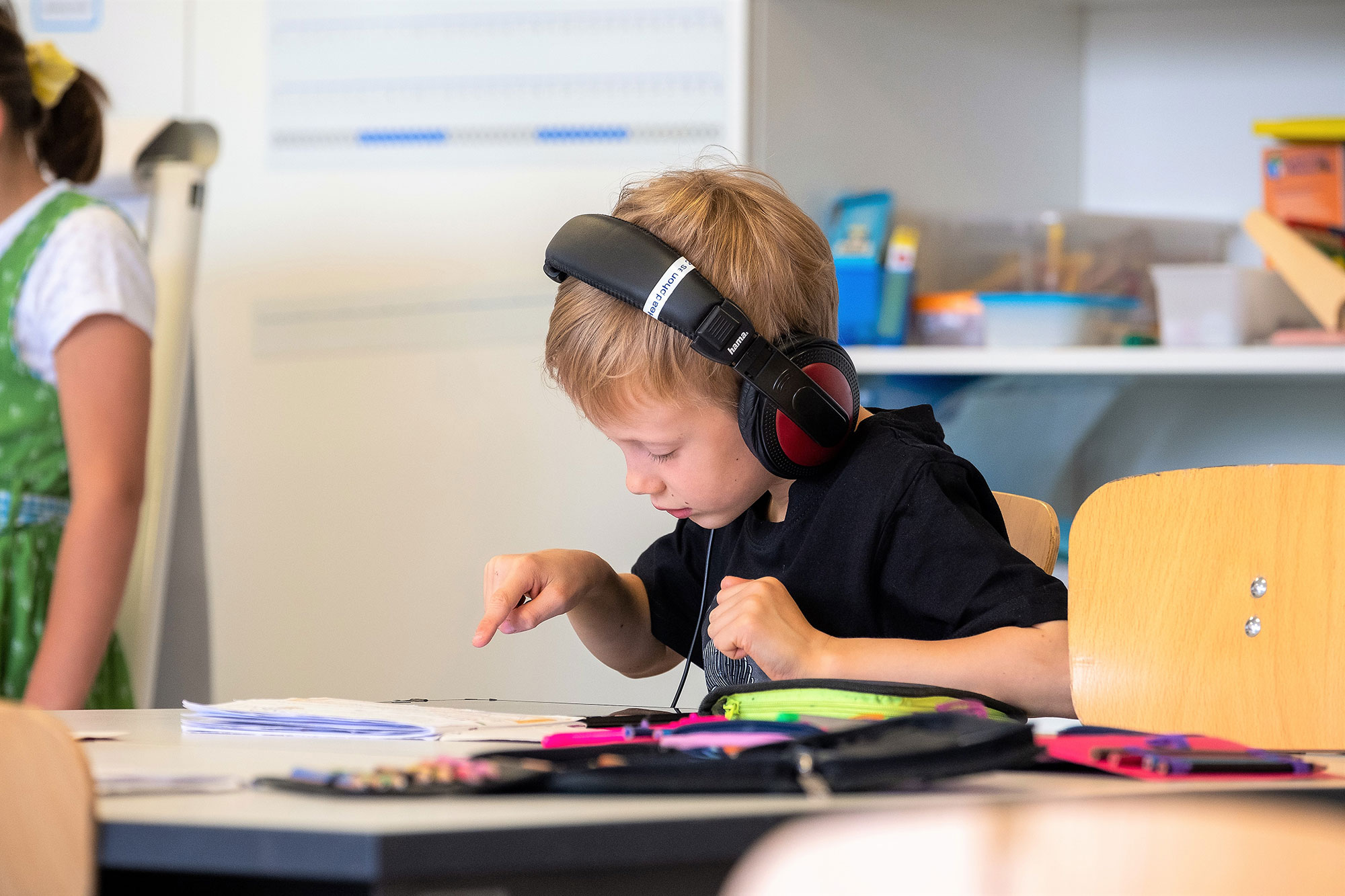 Ein Junge sitzt am Schreibtisch im Klassenzimmer, trägt Kopfhörer, die an ein Tablet verbunden sind und tippt auf den Bildschirm. 	