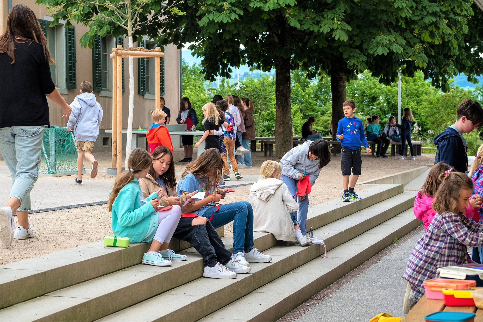 Schülerinnen und Schüler sitzen und spielen draussen auf dem Pausenplatz der Schule mit grossen, grünen Bäumen im Hintergrund. 	