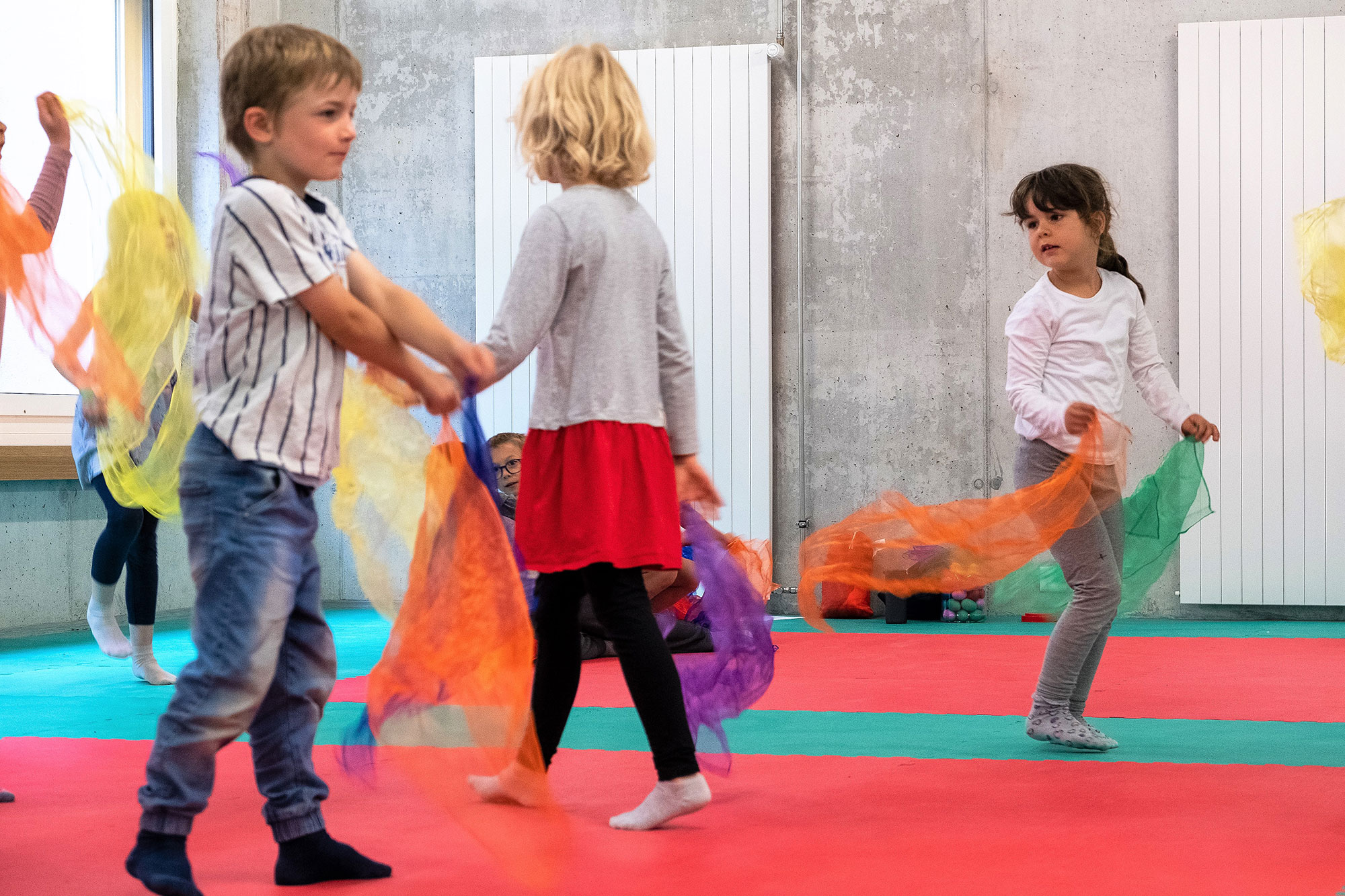 Kinder tanzen und bewegen sich im Sportraum und tragen dabei bunte Seidentücher in den Händen.	