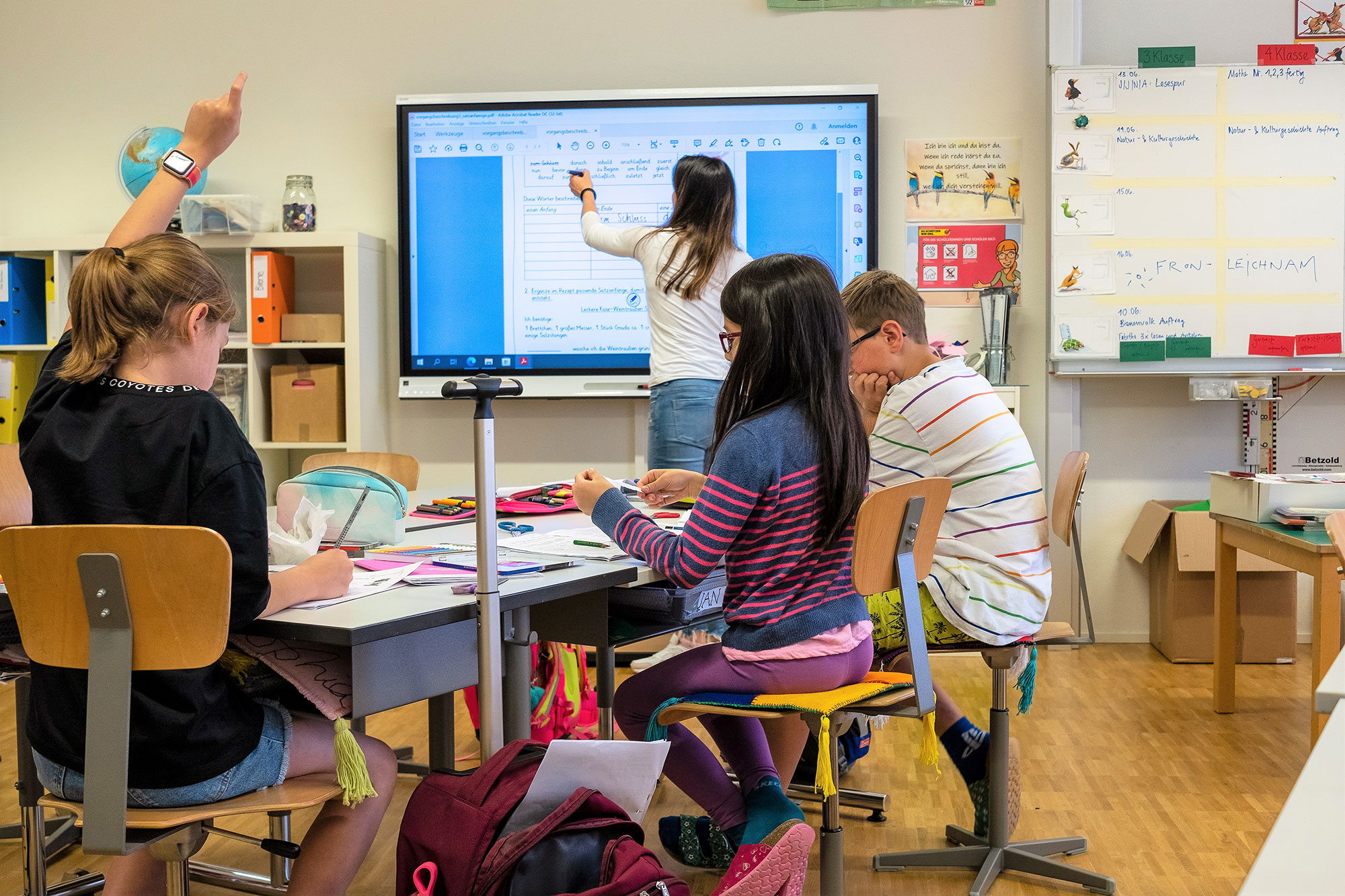 Primarschulkinder sitzen im Unterricht und die Lehrerin steht vorne am Smartboard. Ein Mädchen hält die Hand hoch.	
