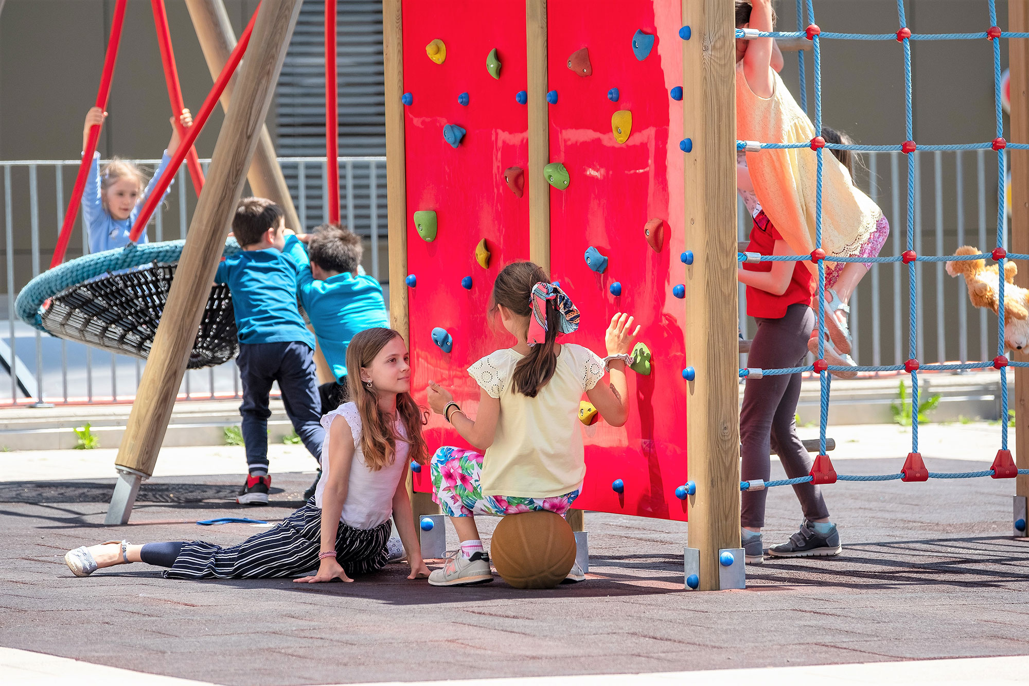 Children are playing in the large playground. Two girls are in focus and are sitting on the ground talking to each other.	