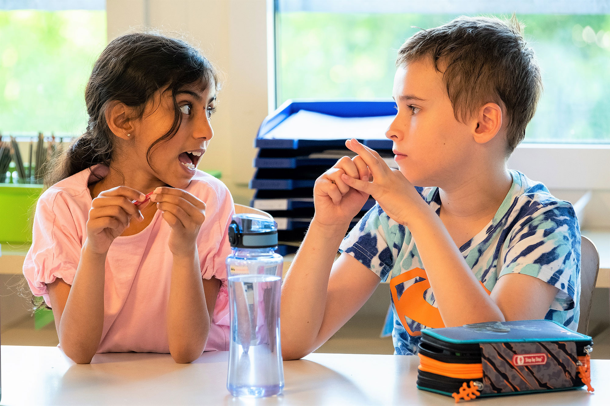 A boy and a girl sit together at the table in a primary school classroom and discuss something.	