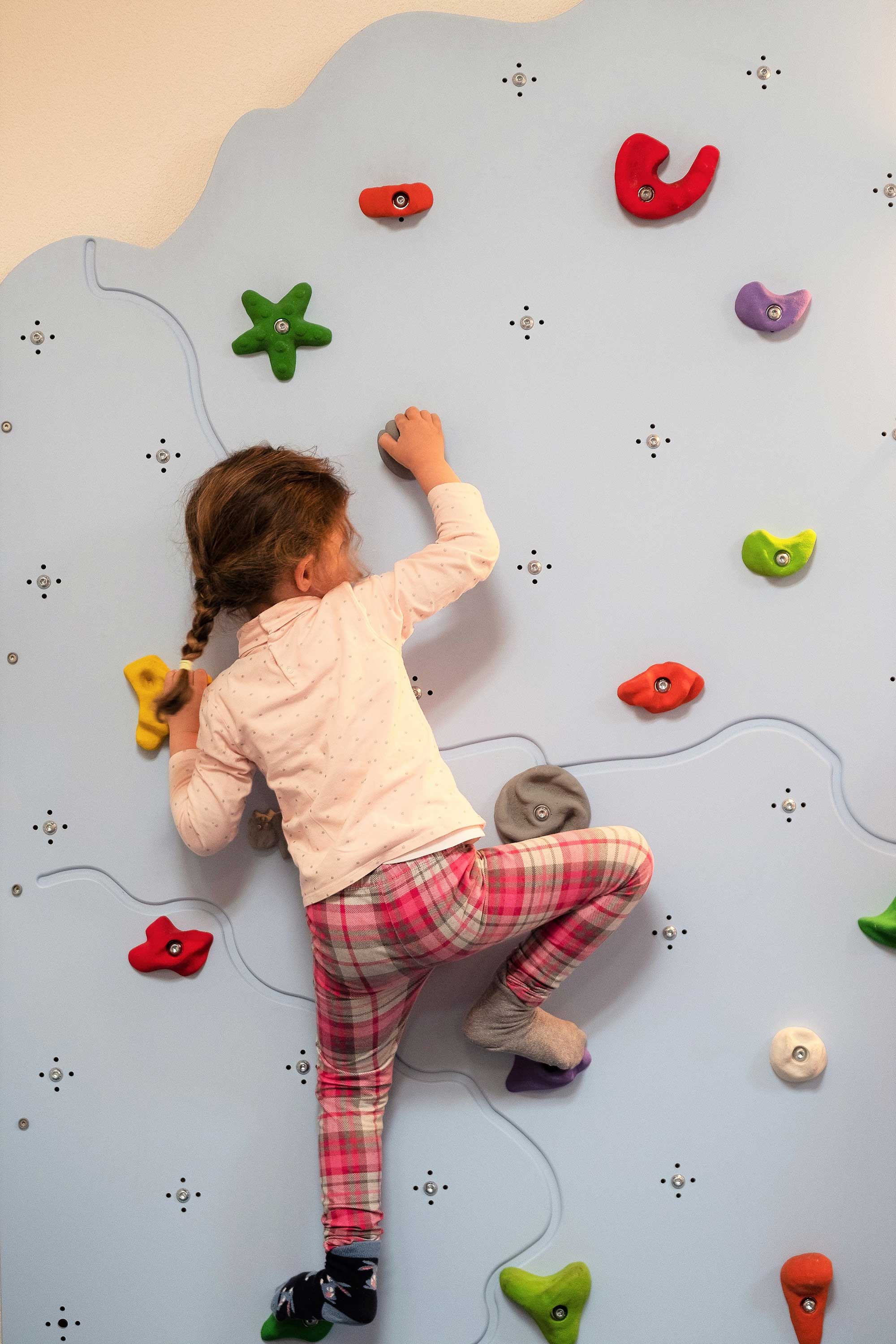 A little girl holds onto the blue climbing wall in the kindergarten.	