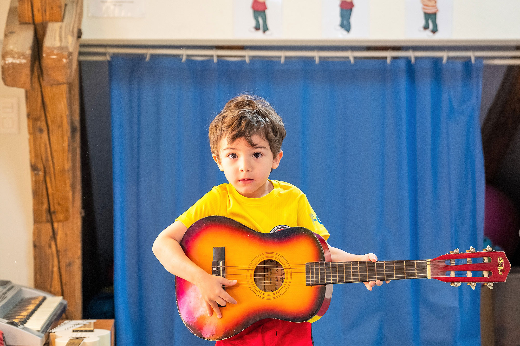 A little boy holds a guitar and looks into the camera with big eyes.	