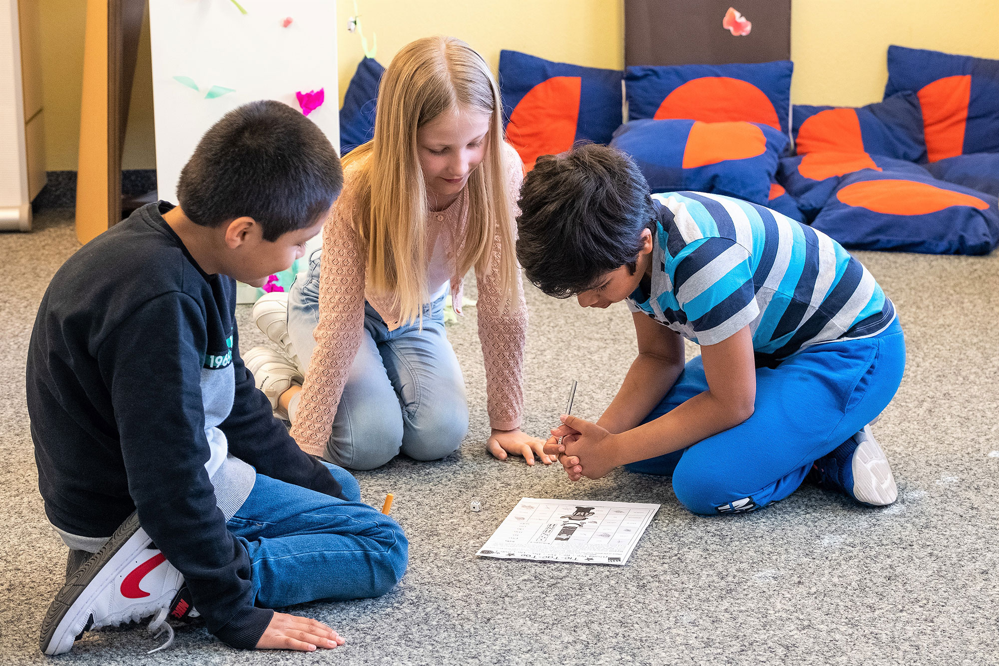 Ein Mädchen und zwei Jungen sitzen auf dem Boden und lernen anhand eines Spiels mit einem Würfel.	