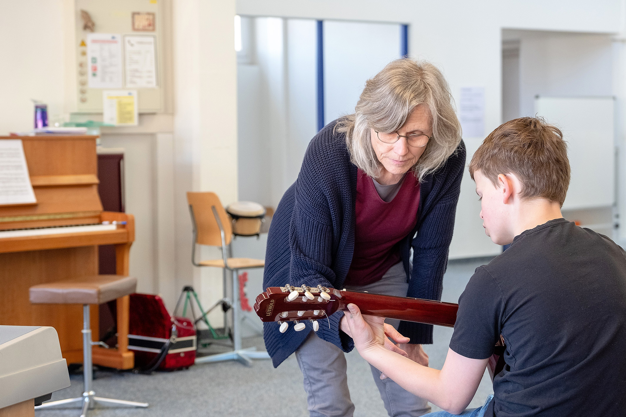 A boy practices playing the guitar and the teacher stands by his side.	