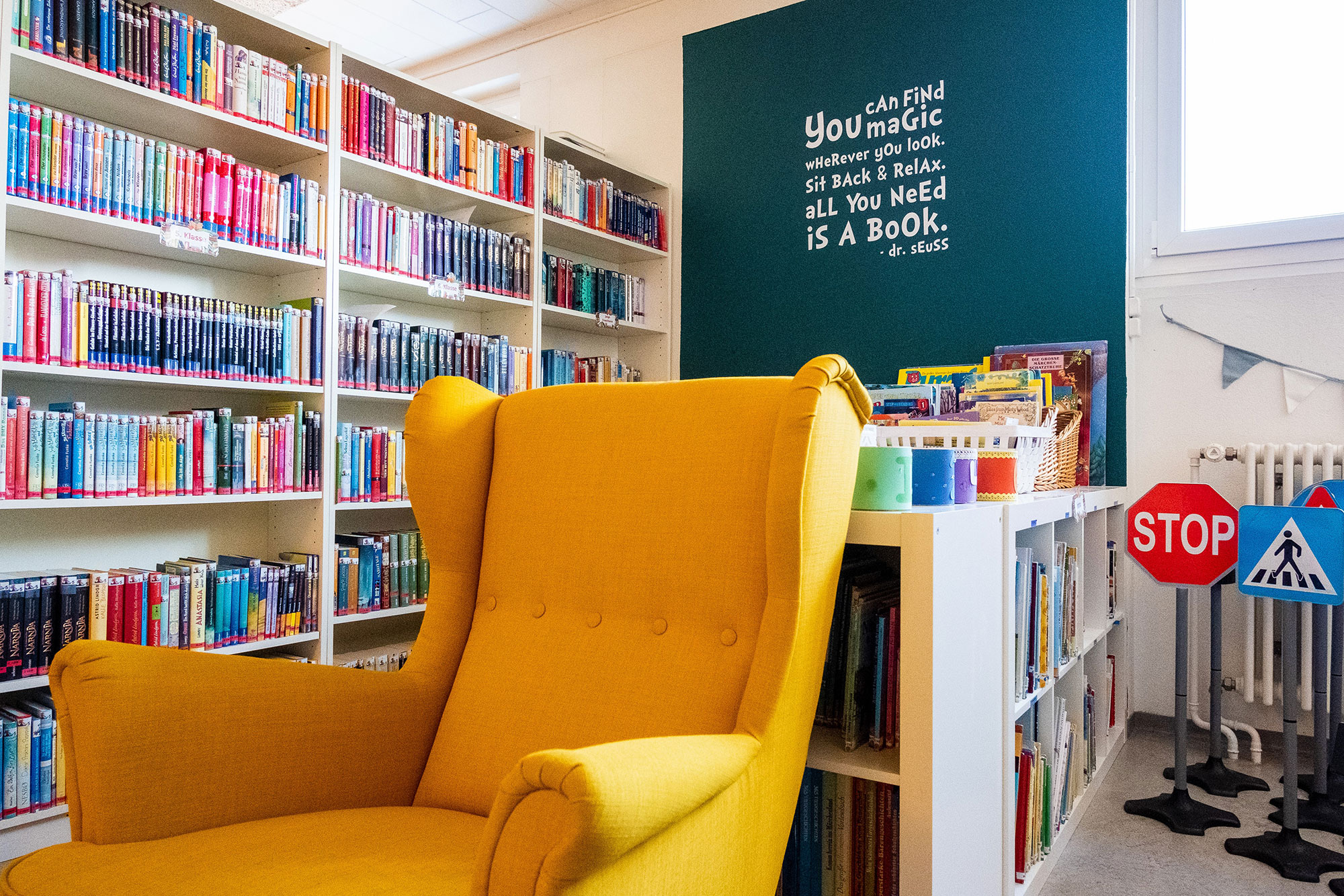 Ein gelber Sessel steht in der farbigen Bibliothek und an der Wand steht der Spruch "You can find magic wherever you look. Sit back and relax. All you need is a book."	