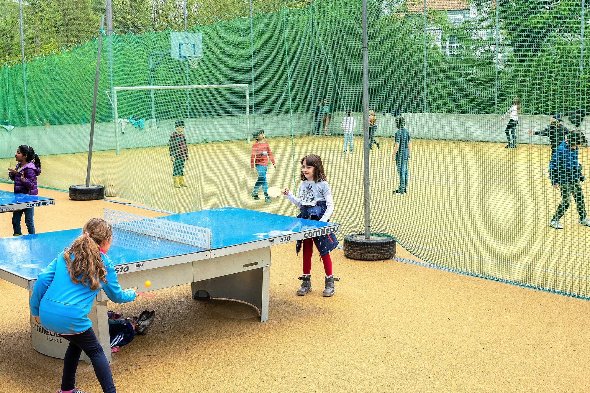 Kinder spielen auf dem grossen Pausenplatz. Eine Gruppe spielt Tischtennis und eine andere Gruppe spielt Fussball.	