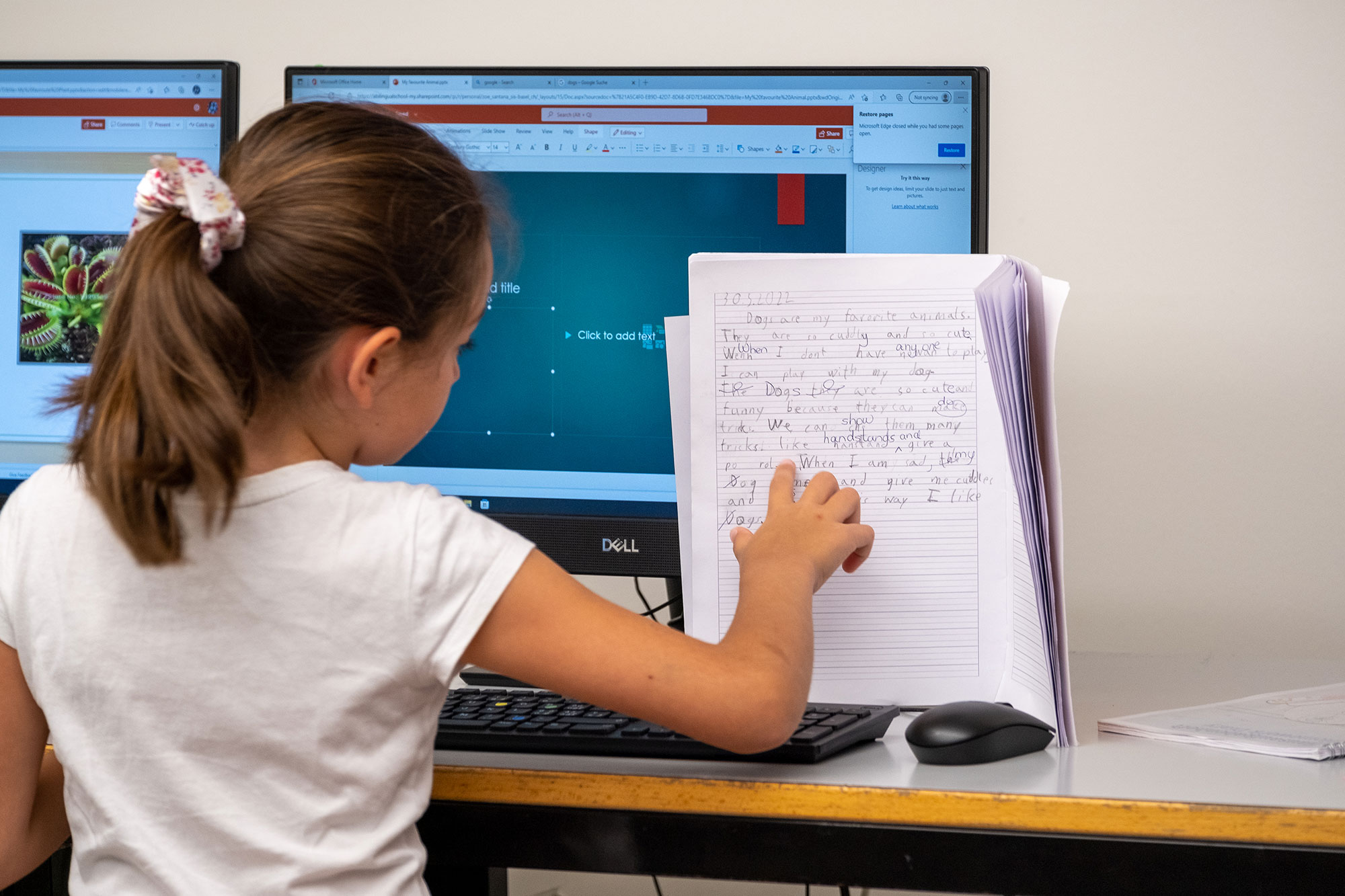 Ein junges Mädchen sitzt vor einem Computer, nebenan hat sie ihr Notizheft. Sie schaut zum Notizheft und zeigt mit dem Finger darauf.	