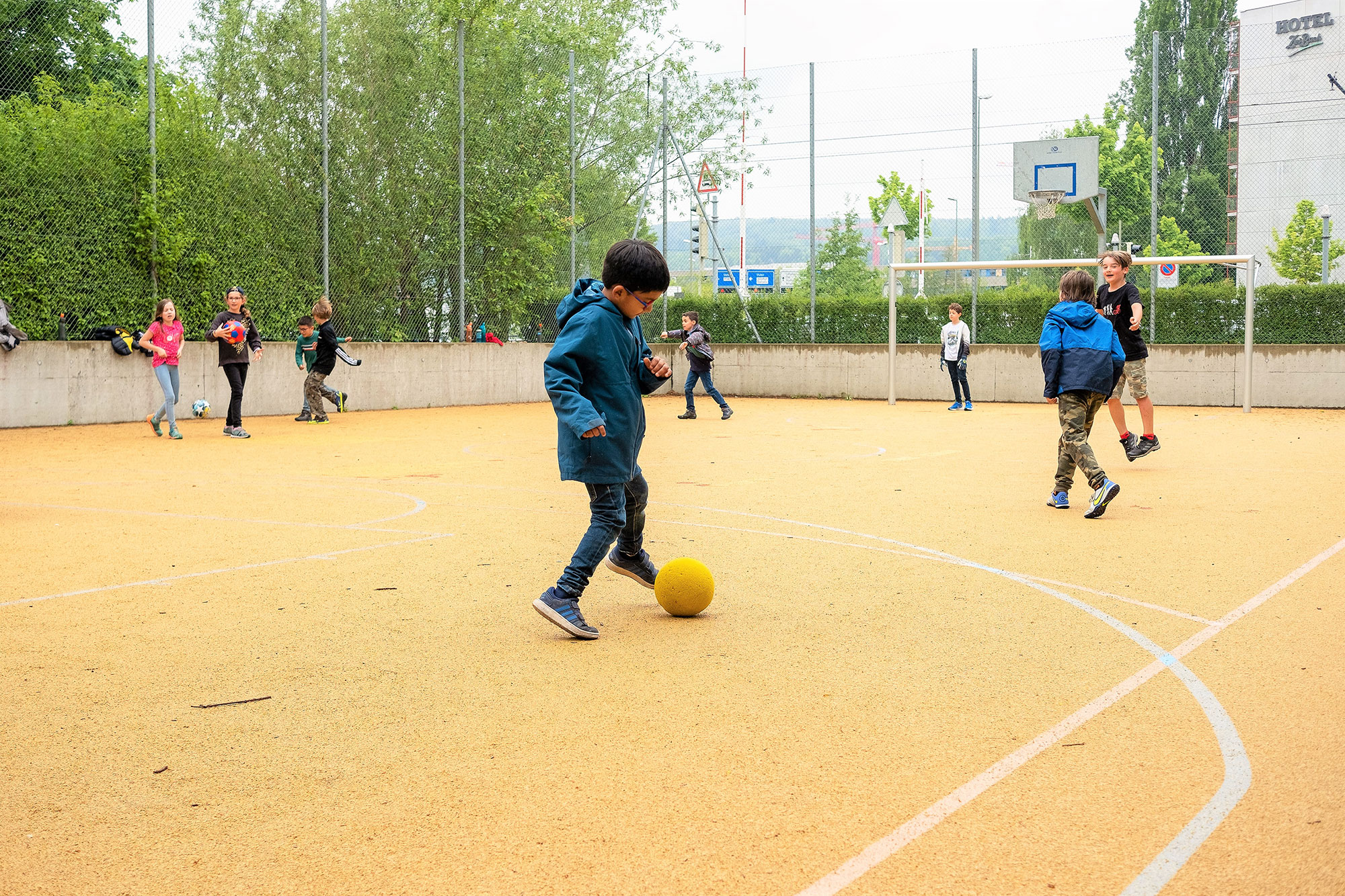 Ein paar Kinder spielen auf dem Sportplatz vor der Schule. Im Fokus ist ein Junge, der mit einem Fussball spielt.	