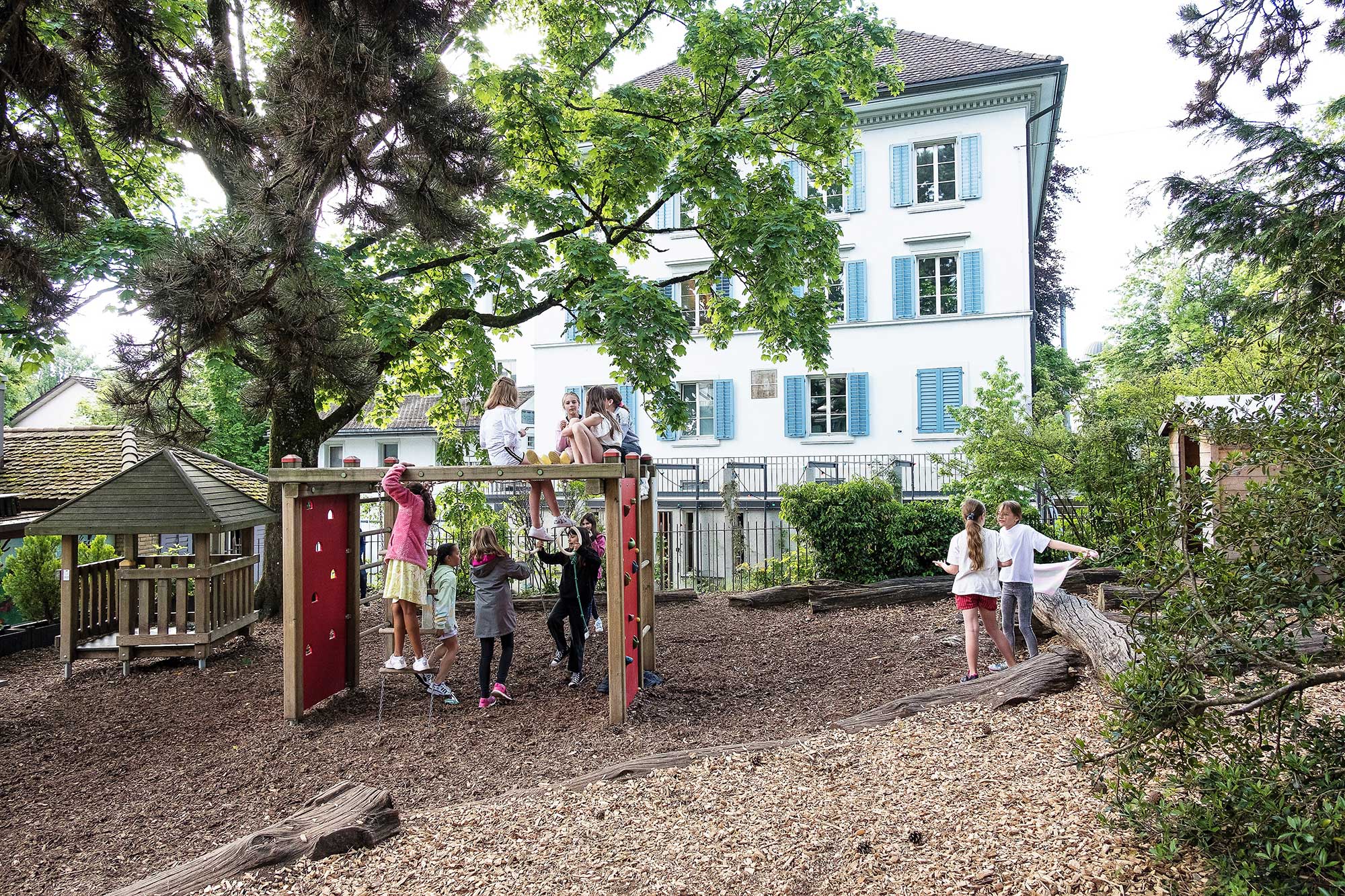 Kinder spielen auf dem Spielplatz der Schule. Im Hintergrund steht ein grosser Baum.	