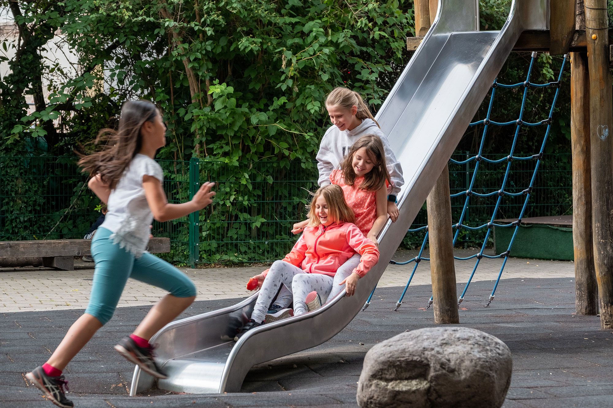 Four primary school girls are outside at the playground. Three girls are on the slide together and one girl is running. 	