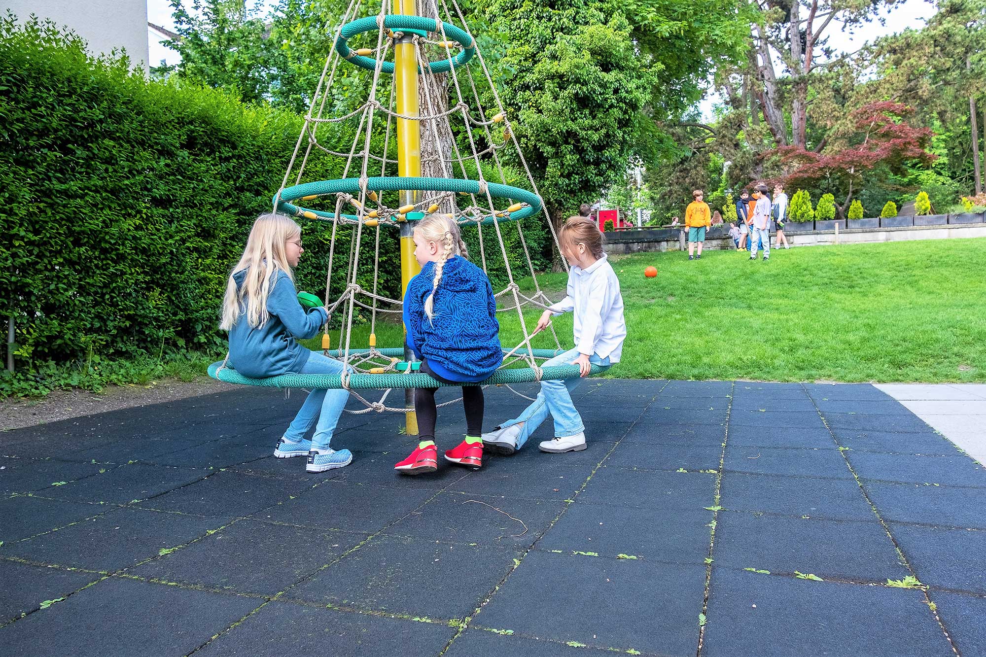 Drei Mädchen sitzen auf dem Karussell auf dem Spielplatz der Schule. Im Hintergrund sieht man eine Jungengruppe mit einem Ball.	