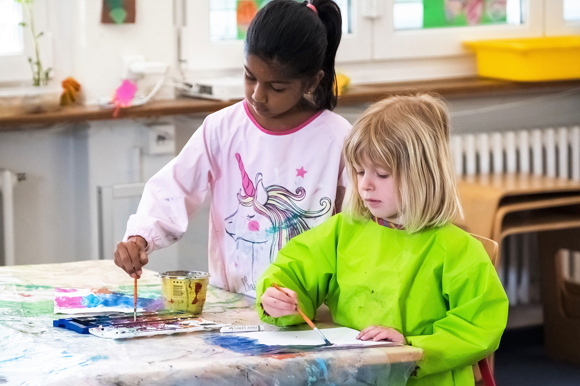 A dark-skinned and a blond girl paint in the kindergarten.	