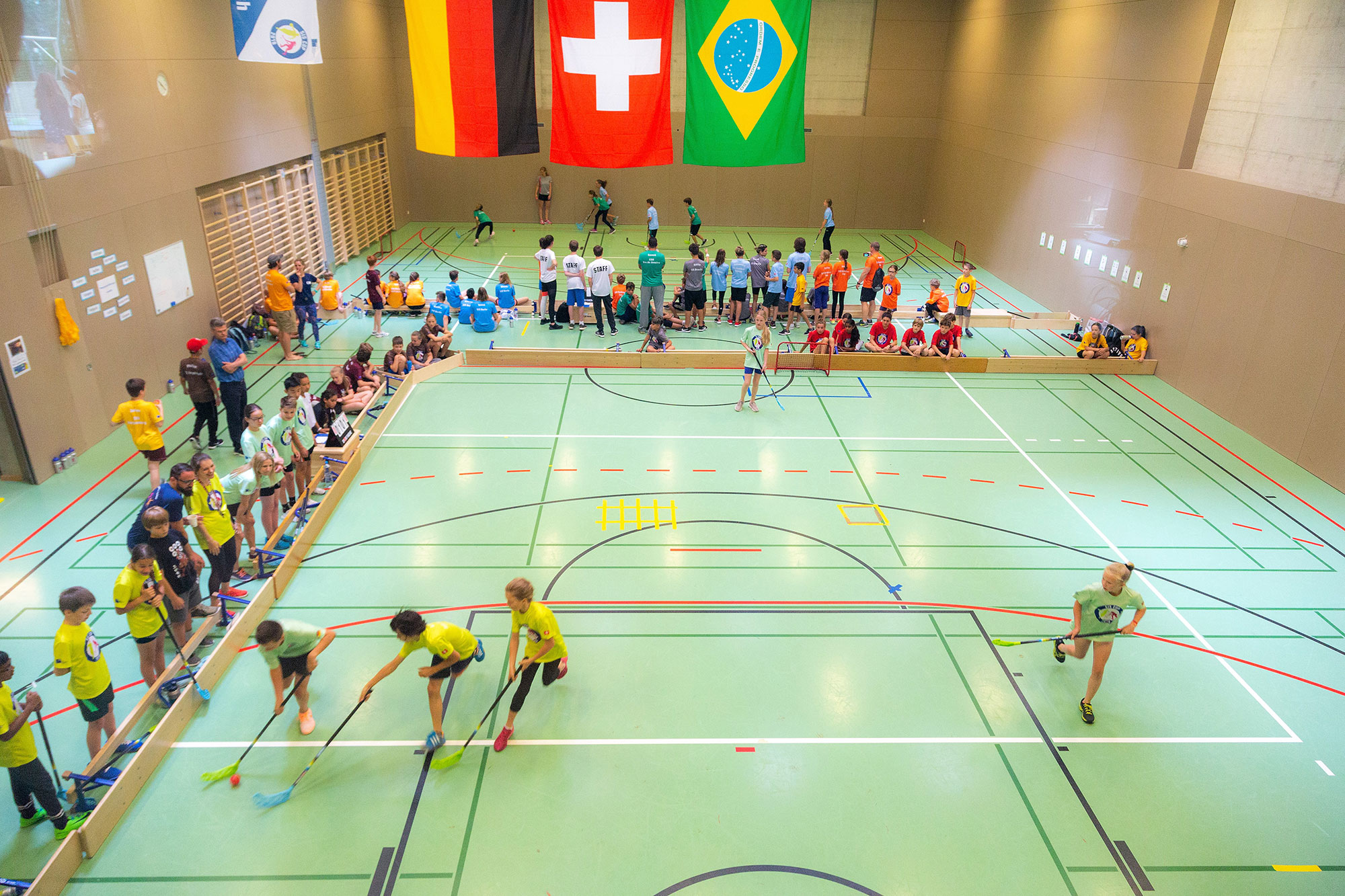 In einer Dreifachturnhalle findet das Unihockeytunier im Rahmen des SIS Cups statt. Es hängen die Länderflaggen von Deutschland, Brasilien und der Schweiz von der Decke.	