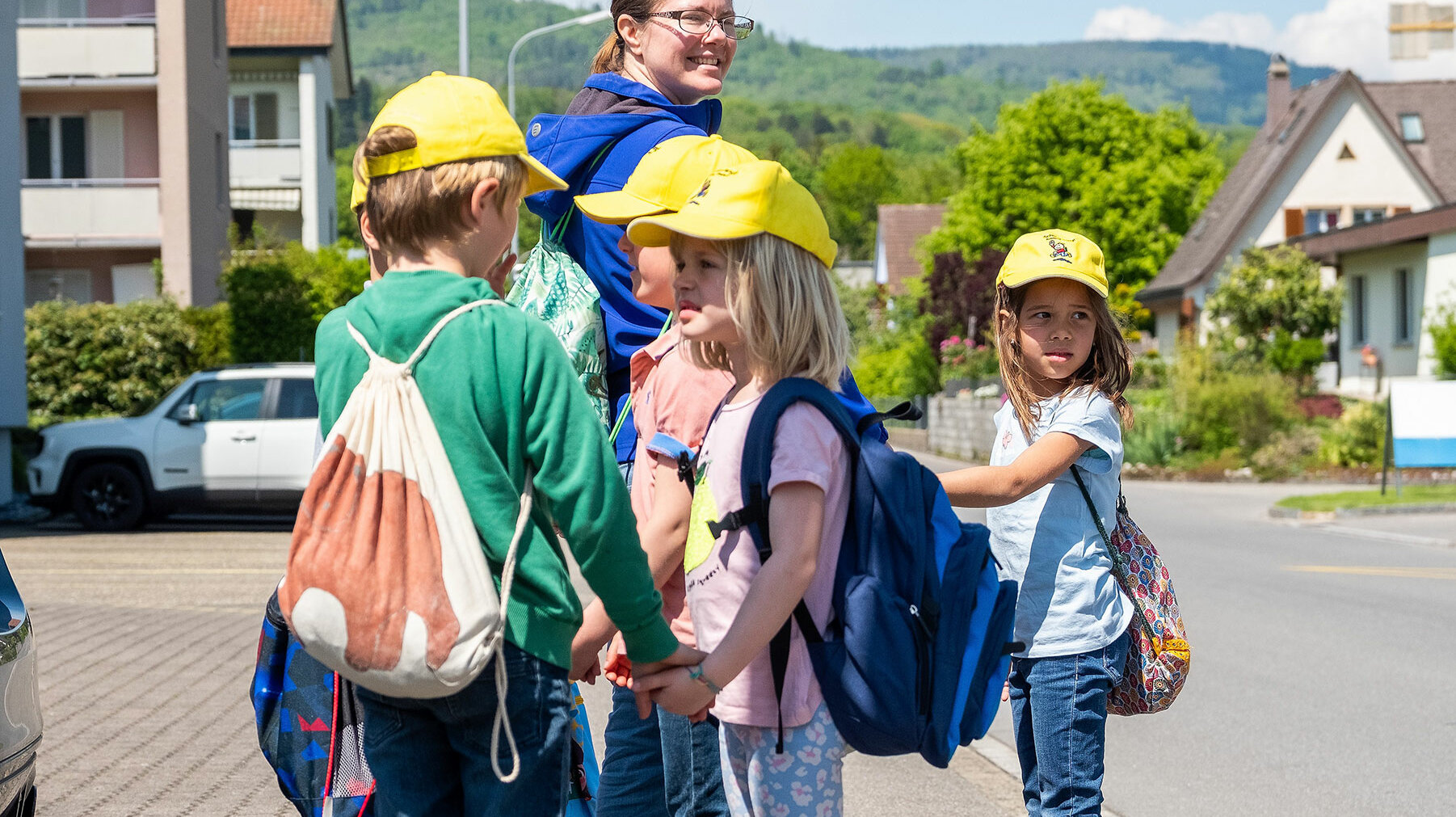 Small children stand in the street behind the teacher and wait. They all wear a yellow cap.	