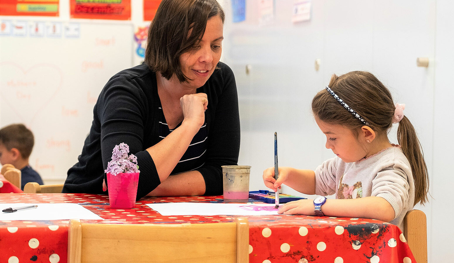 Ein Mädchen malt etwas im Kindergarten. Eine Lehrerin sitzt neben ihr.	