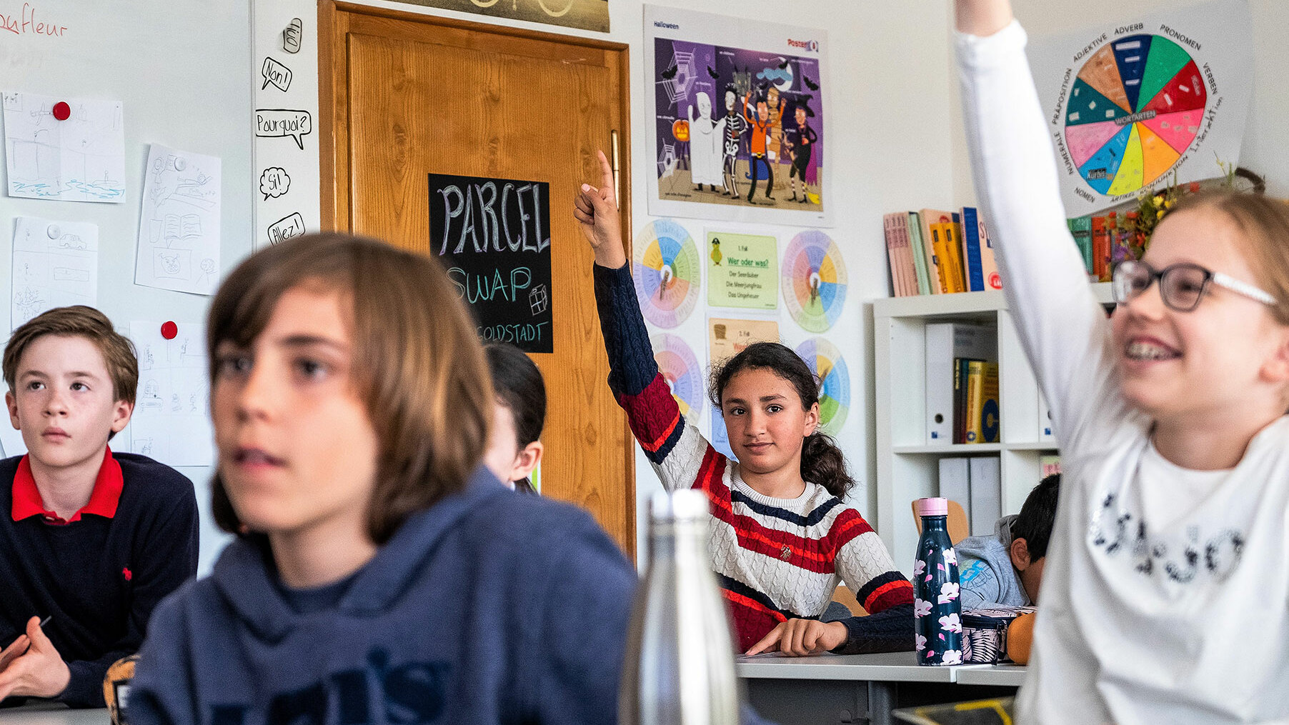 Primarschulkinder sitzen im Unterricht und zwei Mädchen strecken die Hände hoch.	