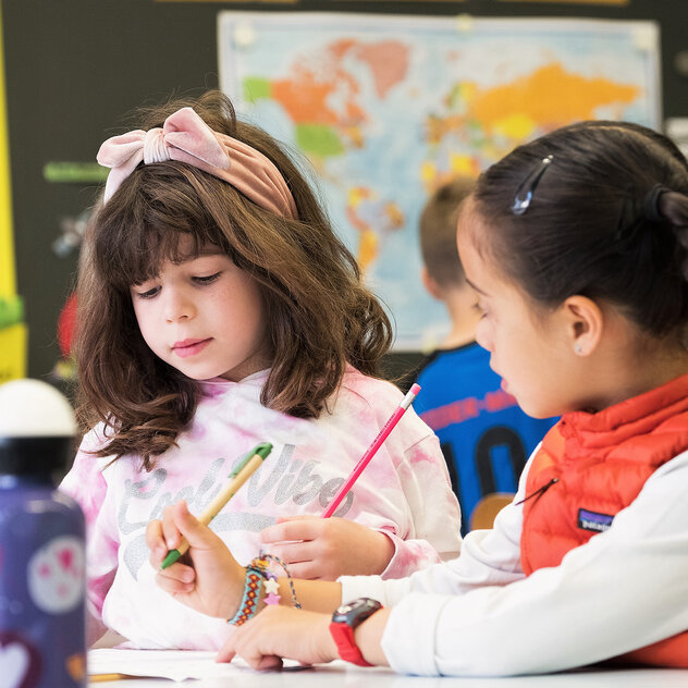 Two primary school girls sit at the desk in class and write something.	