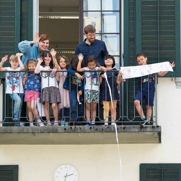 A group of kindergarten children stand on a balcony with two teachers and wave to the audience.