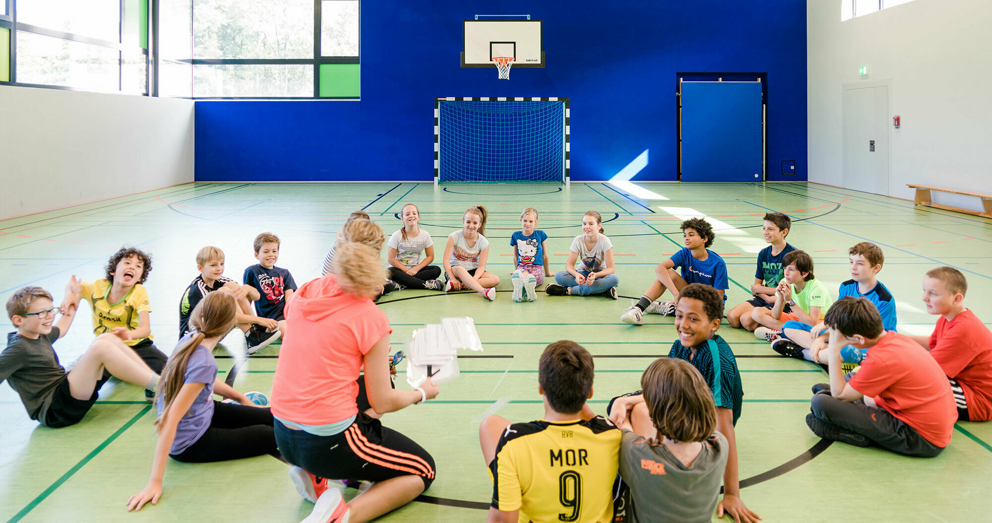 In a modern gymnasium, a sports teacher gathers her students seated in a circle and gives instructions.