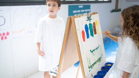 A girl in a painting smock paints coloured elements with a brush on a white sheet of paper at an easel. 