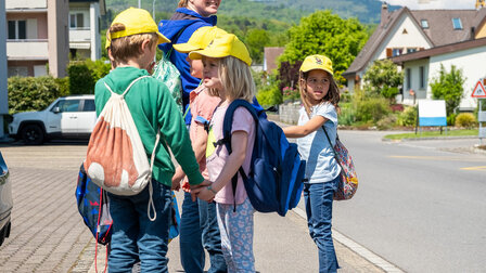 Small children stand in the street behind the teacher and wait. They all wear a yellow cap.	