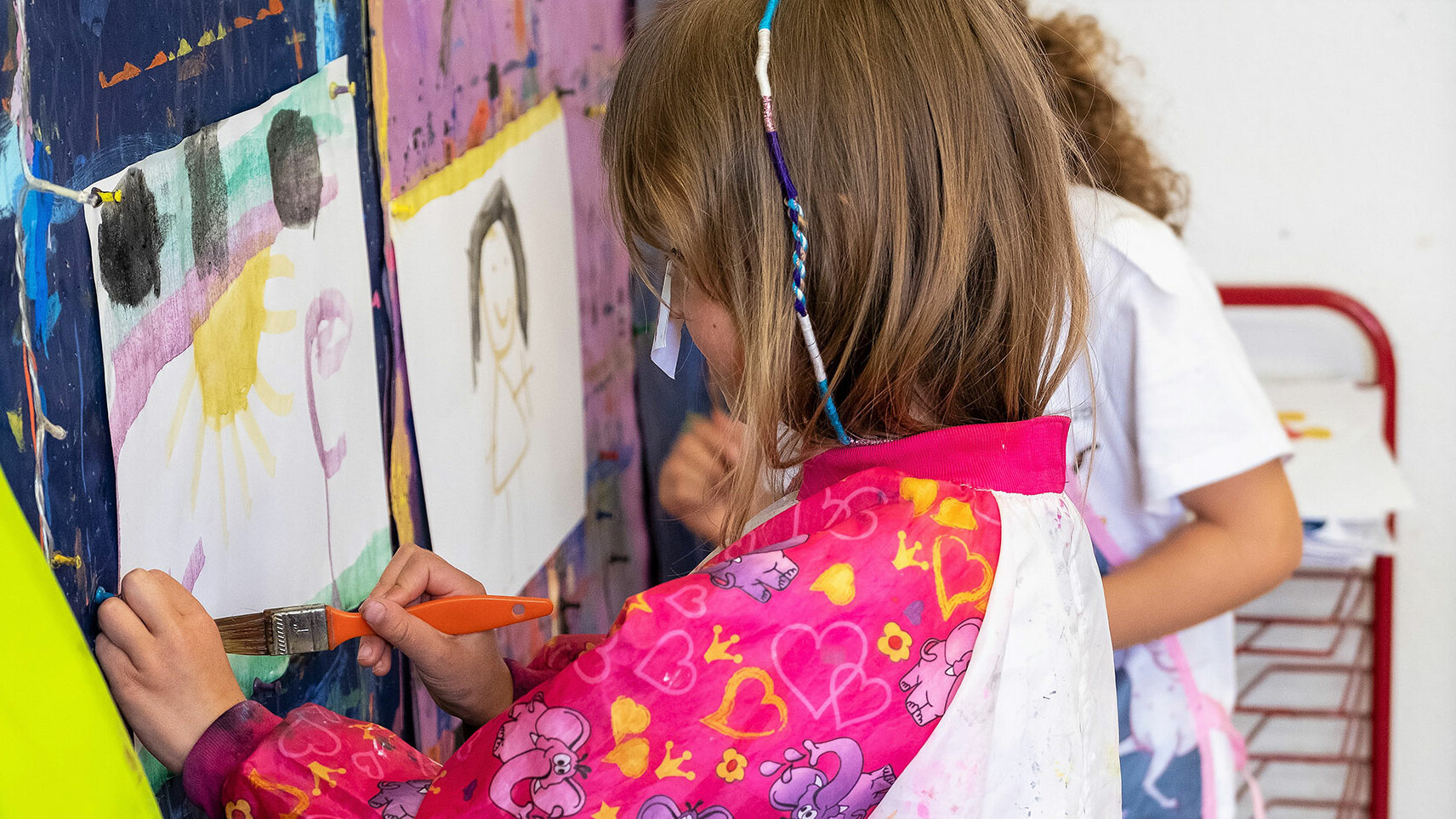 Ein kleines Mädchen aus dem Kindergarten malt ein Bild auf einem Blatt, das an der Wand hängt.	
