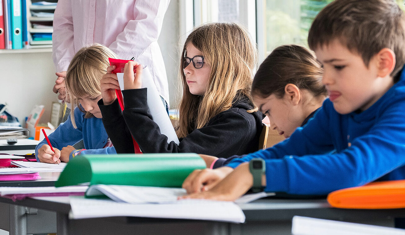 Primarschülerinnen und -schüler arbeiten konzentriert im Schulzimmer während dem Unterricht. Im Hintergrund steht die Lehrerin.