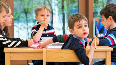 Little boys are sitting at a table and one of the boys is looking back with big blue eyes.	