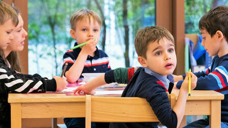 Little boys are sitting at a table and one of the boys is looking back with big blue eyes.	