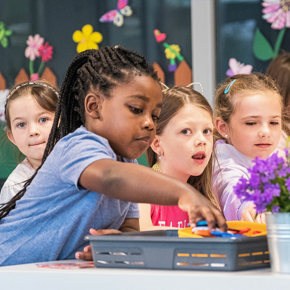 Sechs kleine Mädchen sitzen an einem Tisch. Ein Mädchen greift nach einem Stift aus einer grauen Box. Es sind zwei kleine Blumentöpfe zu sehen.