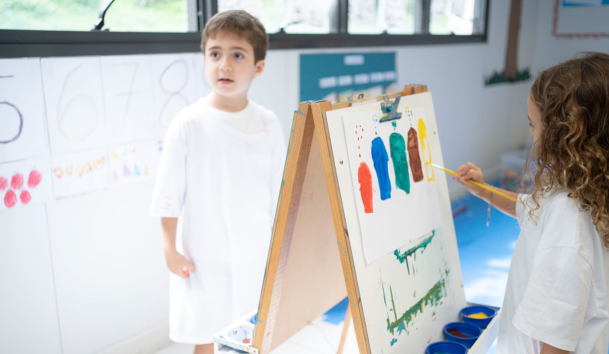 A girl in a painting smock paints coloured elements with a brush on a white sheet of paper at an easel. 