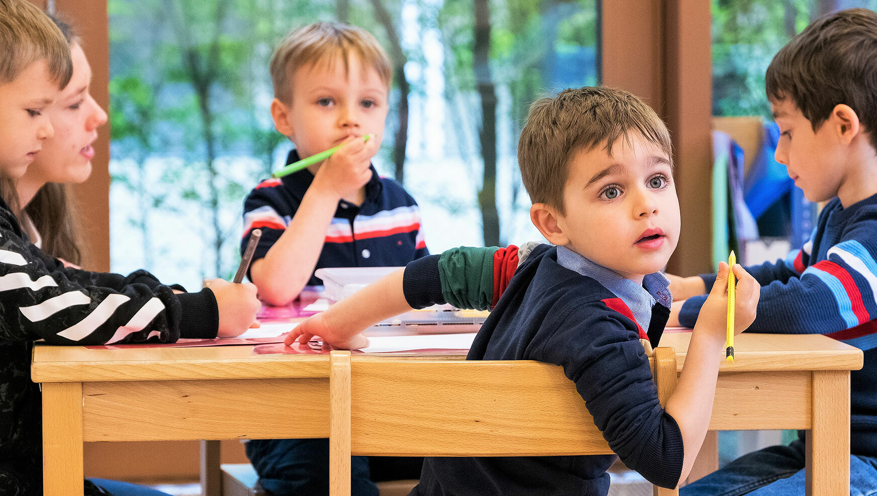 Kleine Jungs sitzen an einem Tisch und einer der Jungs schaut mit grossen blauen Augen nach hinten.	