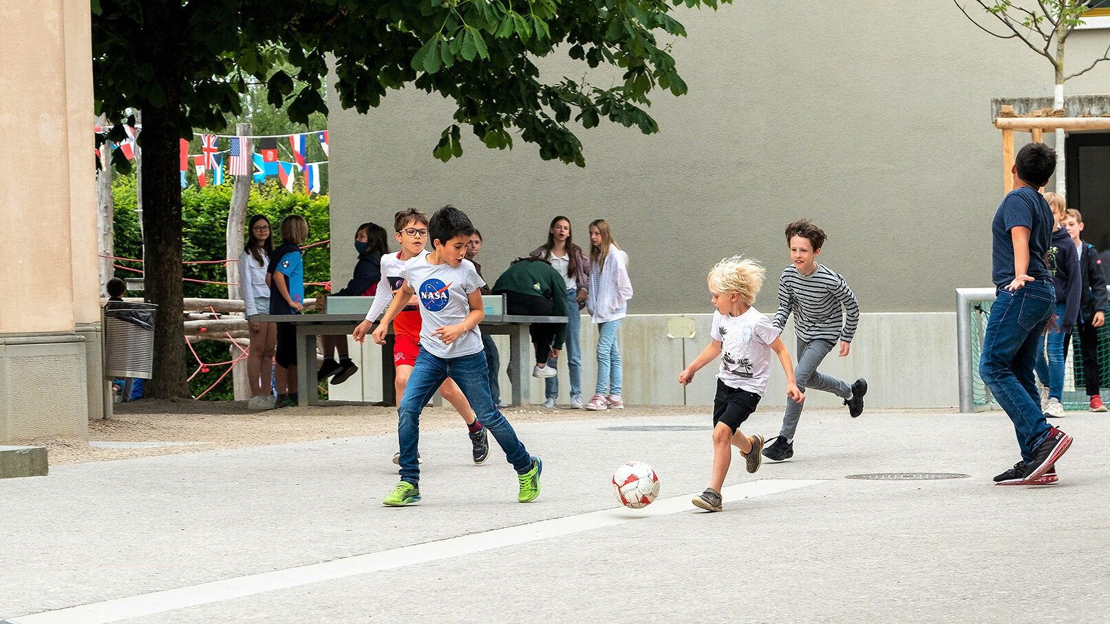 Eine Gruppe Kinder spielen draussen auf dem Pausenplatz der Schule Fussball.	